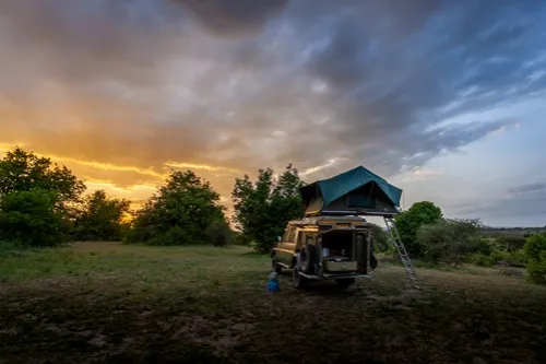 An olive-green 4x4 vehicle with a dark green canvas rooftop tent pitched in a grassy clearing at sunset. The back of the vehicle is open, showing the cooking/storage area. A ladder descends from the tent. The sky is split dramatically, with warm orange and yellow light on the left fading into deep blue and grey storm clouds on the right.