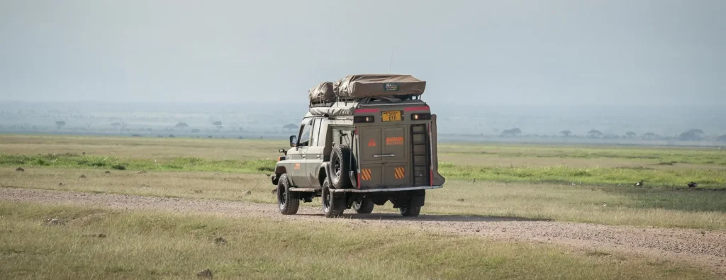 A vehicle with luggage on top drives along a dirt road in Kenya National Parks, surrounded by natural scenery.