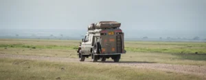 A vehicle with luggage on top drives along a dirt road in Kenya National Parks, surrounded by natural scenery.